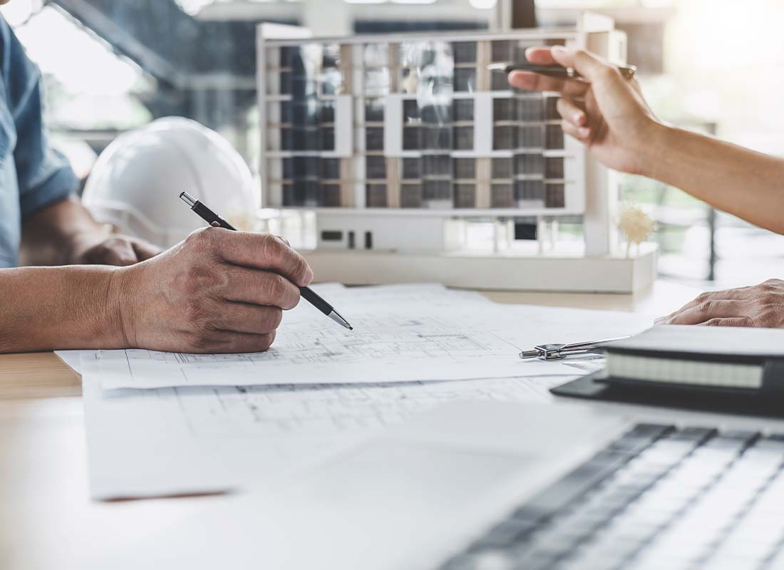 Builders Risk Insurance - Hands of Architect and Engineer Working on a Blueprint with Model Building and Engineering Tools of Working Site in the Background