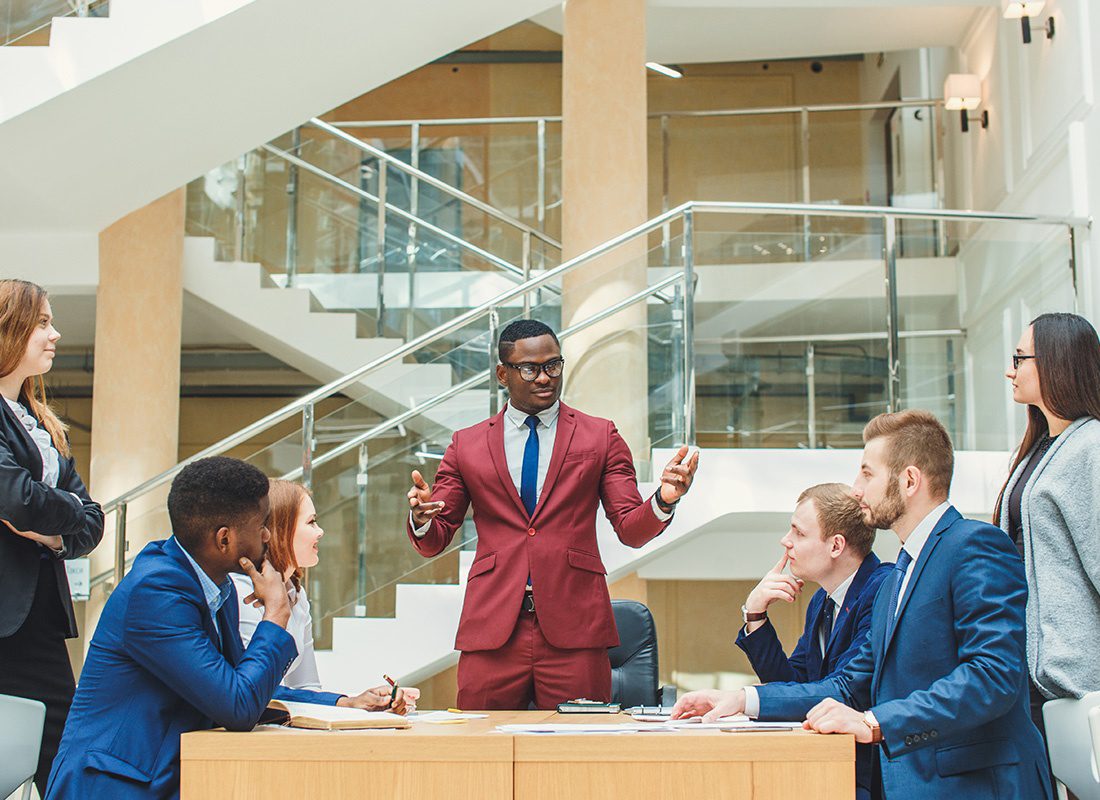 Insurance Solutions - Group of Business Professionals Gathered Together Standing and Sitting Listening to a Man in a Red Suit Speak at an Office